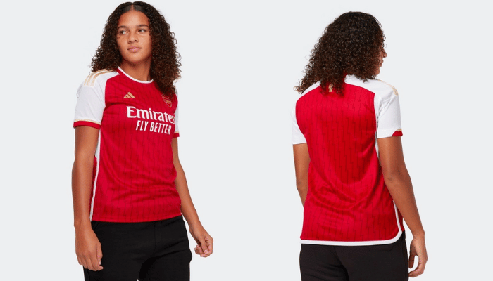 Camisa Titular Feminina do Arsenal Adidas 2023/24 em vermelho, branco e dourado. Detalhes como raios dourados na nuca, faixas laterais com 26 vitórias e 12 empates de 2003-2004, dorsais brancas e gola redonda vermelha com contorno dourado. As três listras douradas nas mangas completam um visual elegante e vitorioso.